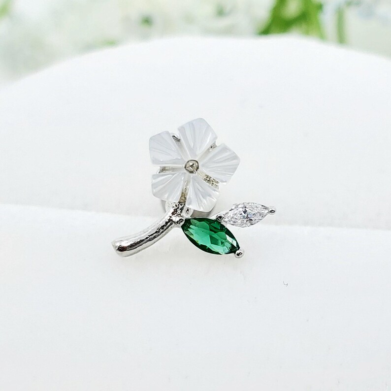May include: A silver-toned metal stud earring shaped like a flower with five white mother-of-pearl petals. The stem features one marquise-cut clear crystal and one marquise-cut green gemstone, both set in silver-toned metal prongs.