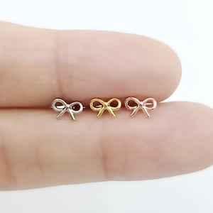 May include: Three small bow-shaped earrings in silver, gold, and rose gold. The earrings are displayed on a light background. The bows are delicate and symmetrical, with a classic design. These earrings are suitable for everyday wear.