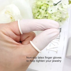 Puede incluir: Dos guantes de dedos de látex blanco para ayudar a apretar las joyas. Los guantes se muestran en un dedo.