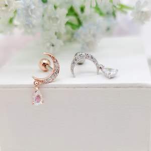 May include: Two earrings featuring a crescent moon design. One is rose gold with clear stones and a pear-shaped iridescent charm. The other is silver with clear stones and a clear pear-shaped charm. The earrings are displayed on a white surface.