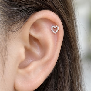 May include: A silver-toned heart-shaped cartilage earring featuring a series of small, clear, sparkling stones set along the perimeter. The jewelry is positioned in the upper ear, showcasing a delicate and refined design with a polished metal finish.
