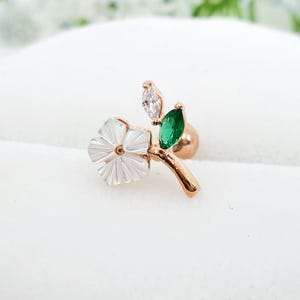 May include: A rose gold tone stud earring featuring a white mother of pearl flower, a marquise cut clear crystal, and a marquise cut green crystal. The design includes a curved stem and a round backing, creating a floral and leaf motif.