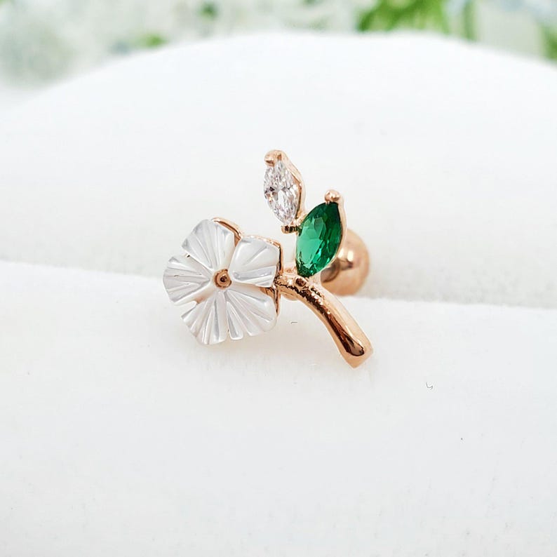 May include: A rose gold tone stud earring featuring a white mother of pearl flower, a marquise cut clear crystal, and a marquise cut green crystal. The design includes a curved stem and a round backing, creating a floral and leaf motif.