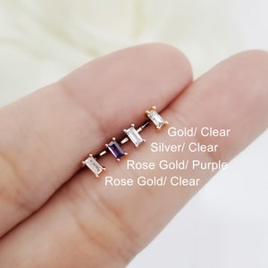 May include: Four different colored earrings with a rectangular clear or purple stone. The earrings are gold, silver, rose gold, and rose gold with a purple stone. The text below the earrings reads "Gold/Clear", "Silver/Clear", "Rose Gold/Purple", and "Rose Gold/Clear".