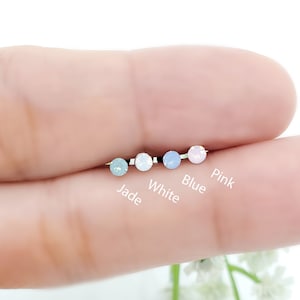 May include: Five small round opal nose rings in different colors: jade, white, blue, and pink. The rings are displayed on a finger.