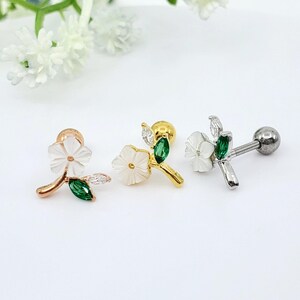 May include: Three stud earrings featuring a white flower design with a green marquise-cut stone leaf and a clear marquise-cut stone leaf. The earrings are available in rose gold, yellow gold, and silver tones, each with a flat back post.