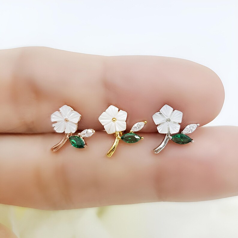 May include: Three flower-shaped stud earrings featuring white mother-of-pearl petals, a central gold-tone dot, and a stem adorned with one marquise-cut green gemstone and one clear marquise-cut gemstone, available in rose gold, gold, and silver tones.