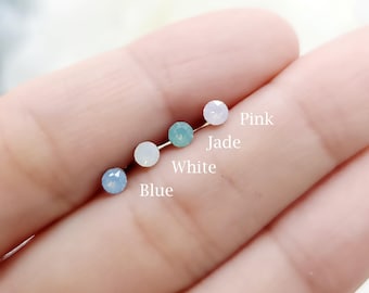 May include: Four small, round, faceted stud earrings arranged in a row, labeled with text: Blue, White, Jade, and Pink. Each earring features a single gemstone in its respective color, showcasing a reflective, multi-faceted surface.