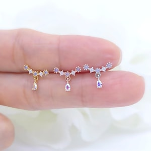 May include: Three delicate earrings are displayed, each featuring a curved bar adorned with small, light purple gemstones. A teardrop-shaped charm dangles from each bar. The earrings are in gold and silver tones, showcasing a minimalist design.