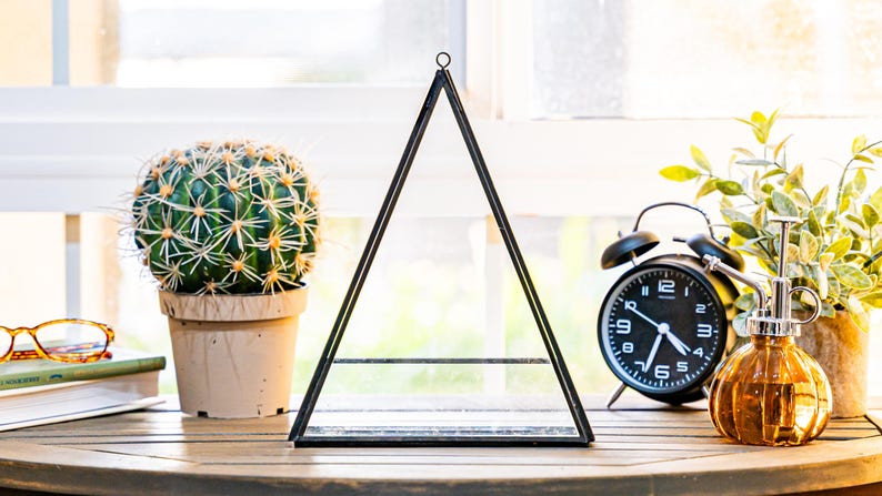 May include: A decorative black metal and glass terrarium in a triangular shape. A potted cactus, a black alarm clock, and a decorative glass bottle are also on the wooden surface. The scene is set near a window.