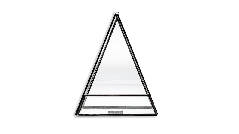 May include: A black metal and glass terrarium in a triangular shape. The terrarium has a small shelf and a loop at the top for hanging. The glass panels are clear, and the frame is black.