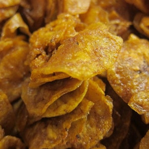 May include: Close-up of a pile of golden-brown banana chips. The chips have a crispy texture and a slightly oily sheen, with visible edges and a natural, irregular shape. The snack appears to be seasoned.