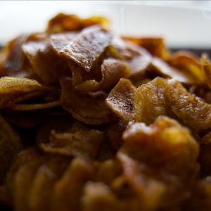 May include: Close-up of a pile of golden-brown, crispy potato chips. The chips have a slightly oily sheen and a textured, irregular surface. The image is well-lit, highlighting the details of the snack.