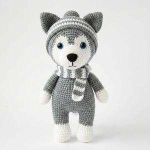 May include: Crocheted husky dog toy wearing a gray and white striped hat and scarf. The dog has blue eyes, a black nose, and white paws. The toy is gray and white and stands upright.