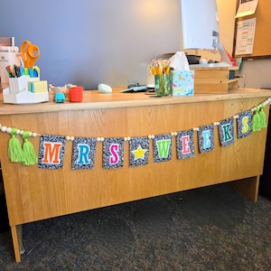 May include: A decorative banner hanging on a wooden desk, featuring black and white speckled composition notebook-style pennants. The letters spell MRS WEEKS in colorful block fonts, with a yellow star between the words and lime green tassels.