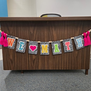May include: A decorative banner featuring seven rectangular pennants with a black and white composition notebook pattern. The pennants display the text WE, a pink heart, and MATH in colourful block letters, accented by wooden beads and pink tassels.