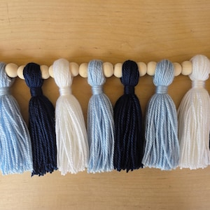 Handmade tassel garland featuring alternating navy blue, Carolina blue, and white yarn tassels strung on a wooden bead garland, displayed on a light wood background