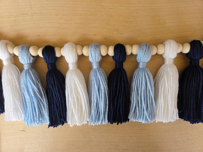 Handmade tassel garland featuring alternating navy blue, Carolina blue, and white yarn tassels strung on a wooden bead garland, displayed on a light wood background