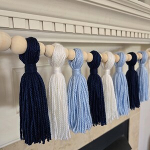 May include: A garland of navy blue, white, and light blue yarn tassels strung on a wooden bead strand. The garland is hanging on a white mantel.