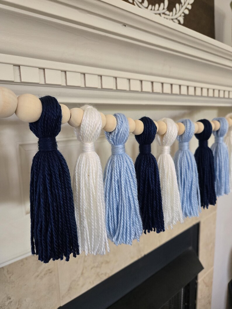 May include: A garland of navy blue, white, and light blue yarn tassels strung on a wooden bead strand. The garland is hanging on a white mantel.
