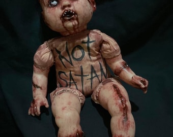 May include: A creepy, bloody baby doll with a stitched-up body and a crown of thorns. The doll is wearing a pink dress with the words "NOT SATAN" written on the front.