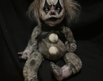 May include: A creepy clown doll with a painted face, wearing a black bow tie and a patterned hat. The doll has a white ball on top of its hat and two white buttons on its chest. The doll's skin is painted in shades of gray and white.