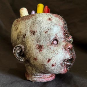 Może przedstawiać: Groteskowy pojemnik na długopisy w kształcie głowy dziecka zombie. Głowa pomalowana na szaro i czerwono, z rozkładającym się wyglądem i widocznymi ranami. Kilka długopisów w różnych kolorach jest włożonych w górną część głowy.