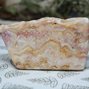 May include: A close-up of a rough, natural, pink and brown banded agate stone. The stone has a layered, wavy pattern and is a light beige color with pink and brown streaks.