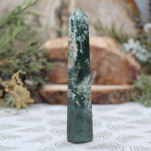 Puede incluir: Una punta de cristal de ágata verde y blanca, también conocida como varita u obelisco, con una superficie lisa y pulida. El cristal mide aproximadamente 10 cm de alto y tiene una forma natural y orgánica.