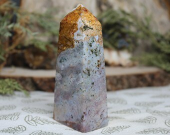 May include: A polished, multi-colored agate crystal point with a golden top. The crystal is a light purple, white, and brown color with a rough, natural texture.