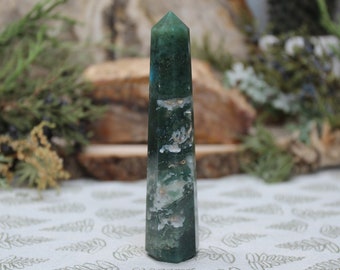 May include: A green and white agate crystal point, also known as a wand or obelisk, is a polished, pointed stone. The crystal is a light green color with white streaks and swirls throughout.