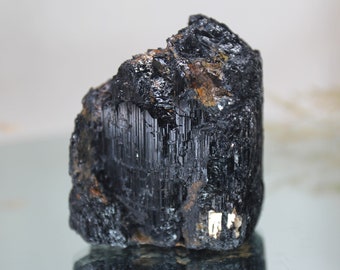 May include: A black, rough-textured, natural crystal with a striated surface. The crystal has a dark, opaque appearance and is a raw, unpolished specimen.