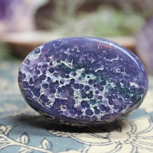 May include: A purple and green agate palm stone with a unique, speckled pattern. The stone is smooth and polished, making it ideal for holding or using in crystal healing.