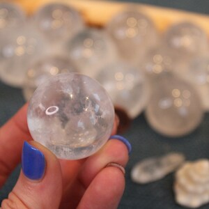 May include: A close-up of a clear quartz crystal sphere, held in a hand with blue nail polish. The sphere is smooth and translucent, with a slight shimmer. The background is blurred, showing other quartz spheres.