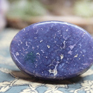May include: A smooth, oval-shaped purple agate stone with a unique, speckled pattern. The stone has a light purple hue with darker purple and white speckles throughout.