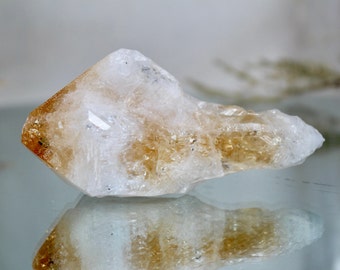 May include: A large, clear, and translucent crystal with a golden yellow hue. The crystal has a pointed tip and a rough, textured surface.