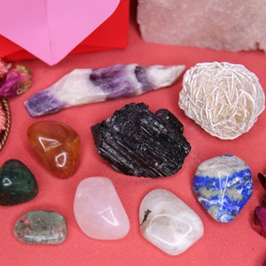 May include: A collection of various crystals and stones, including amethyst, rose quartz, clear quartz, carnelian, black tourmaline, and lapis lazuli. The crystals are arranged on a red surface with dried rose petals and other floral elements.