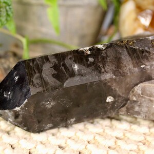 Pode incluir: Um grande cristal de quartzo fumê escuro com uma ponta pontiaguda e uma textura natural e áspera. O cristal é transparente e tem um leve tom fumê. Está descansando sobre uma superfície branca texturizada.