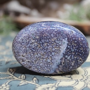May include: A polished, oval-shaped stone with a speckled pattern in shades of blue, white, and tan. The stone has a smooth, glossy surface and appears to be a natural mineral specimen. The overall aesthetic is organic and earthy.