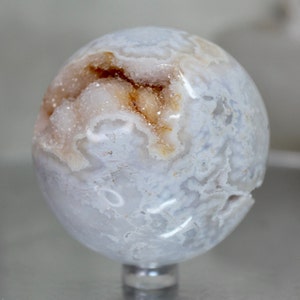 May include: A white agate sphere with a natural crystal formation inside. The sphere has a light blue and white pattern on the outside.