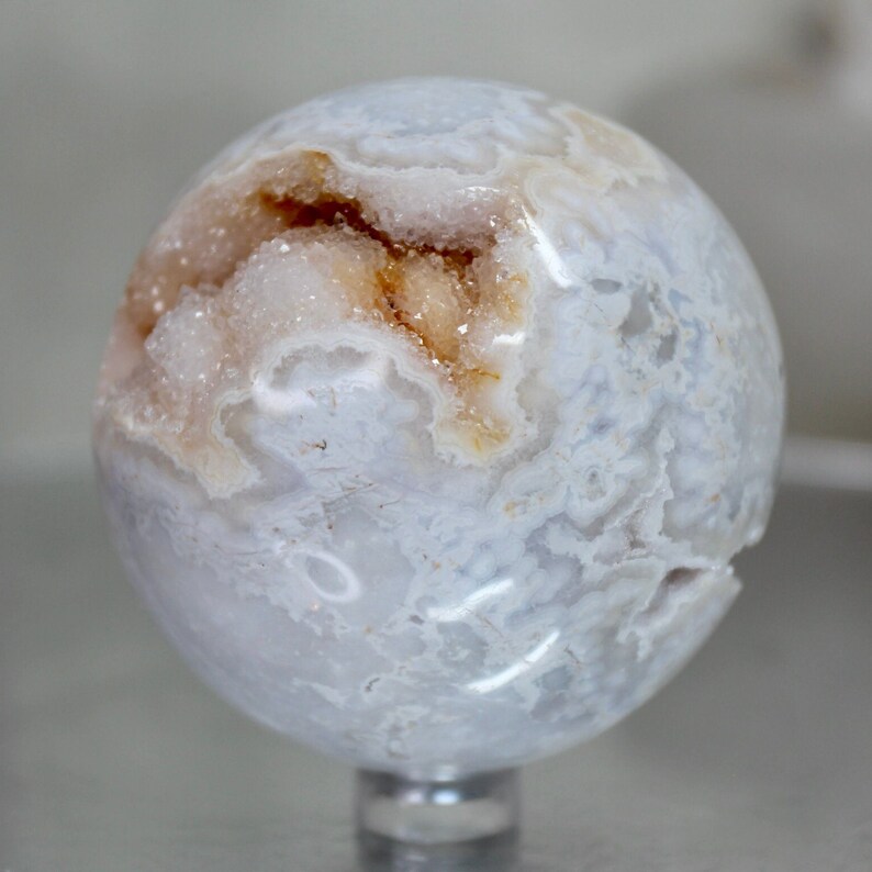May include: A white agate sphere with a natural crystal formation inside. The sphere has a light blue and white pattern on the outside.