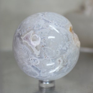 May include: A white agate sphere with a swirling pattern of white and grey. The sphere is polished and has a smooth surface. It is sitting on a clear stand.