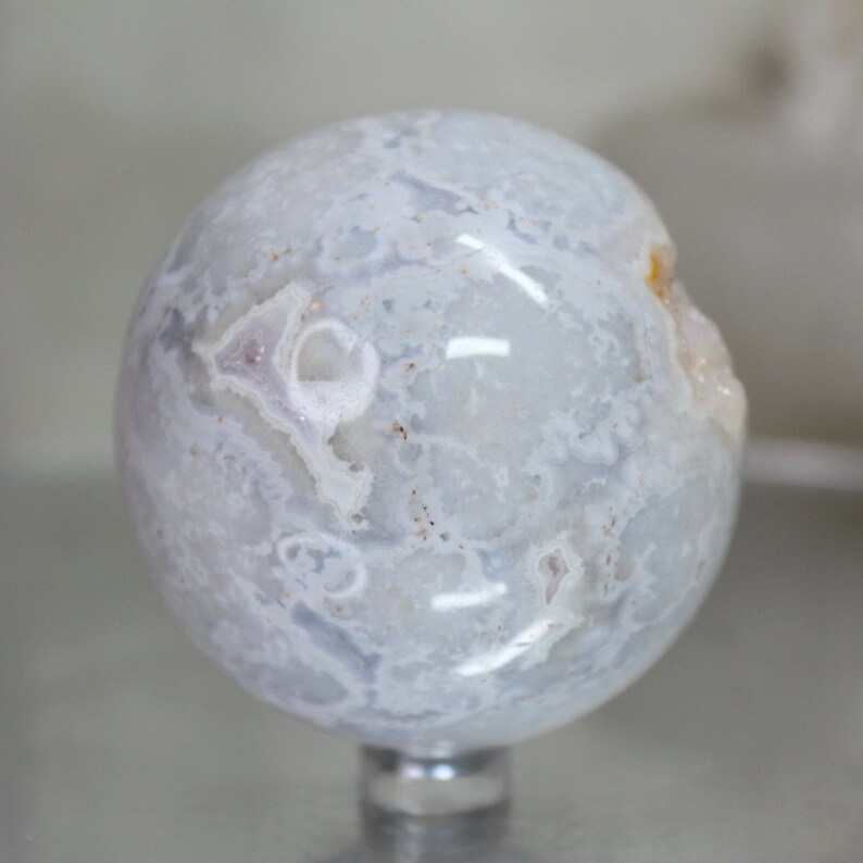 May include: A white agate sphere with a swirling pattern of white and grey. The sphere is polished and has a smooth surface. It is sitting on a clear stand.