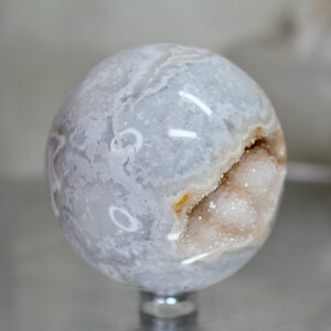 May include: A white agate sphere with a natural crystal druzy opening. The sphere has a light blue and white banded pattern.