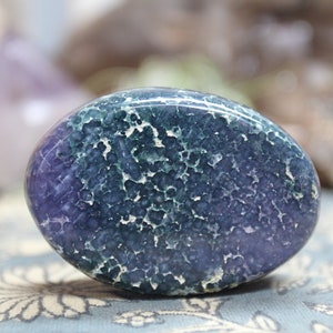 May include: A smooth, oval-shaped polished stone with a swirling pattern of green, purple, and white. The stone has a natural, organic look.