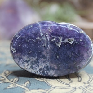 May include: A smooth, oval-shaped purple amethyst stone with white inclusions. The stone is polished and has a glossy finish.