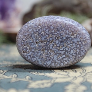 May include: A polished, oval-shaped, light purple and white agate stone with a unique, speckled pattern. The stone has a smooth, polished surface.