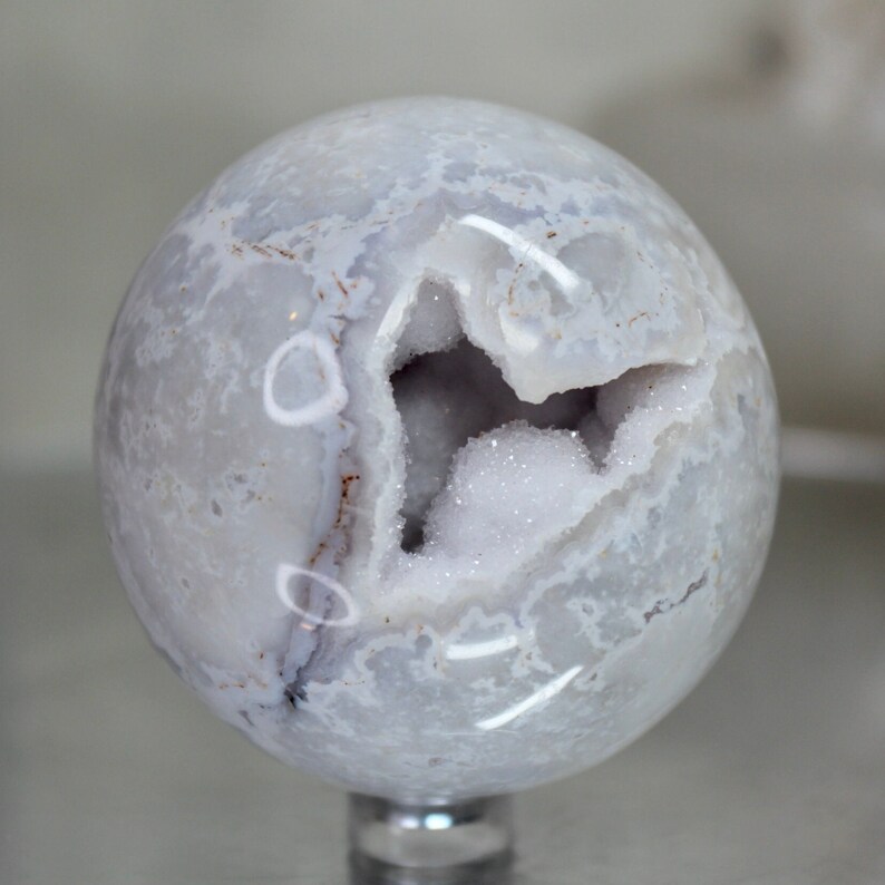 May include: A white agate sphere with a large, open geode cavity. The geode is lined with sparkling white crystals. The sphere is polished and has a smooth, glassy finish.