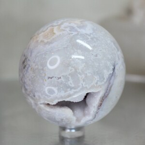 May include: A white agate sphere with a natural crystal formation. The sphere has a smooth, polished surface and a unique, swirling pattern of white and grey.