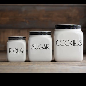 May include: Three white ceramic canisters with black lids and black lettering. The canisters are labeled "Flour", "Sugar", and "Cookies".